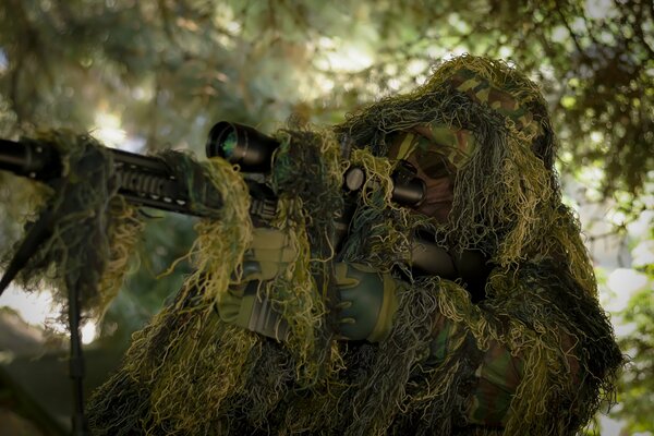 A sniper with a rifle disguises himself in the greenery