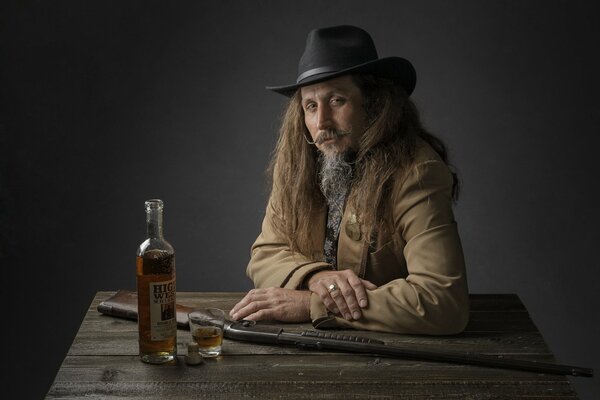 Un uomo dai capelli lunghi con la barba che indossa un cappello da cowboy si siede a un tavolo di legno su cui si trova una bottiglia di whisky e un bicchiere mezzo pieno, e giace anche una pistola