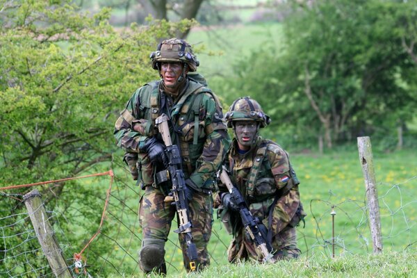 Zwei Soldaten mit Waffen bewegen sich auf einem grünen Feld zwischen Bäumen