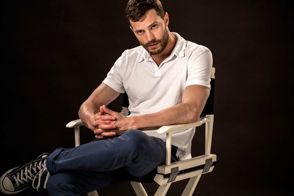 Actor, model and musician Jamie Dornan in a white T-shirt, blue trousers and black sneakers sits in a wooden chair with his fingers crossed and his left leg crossed over his right knee on a black background