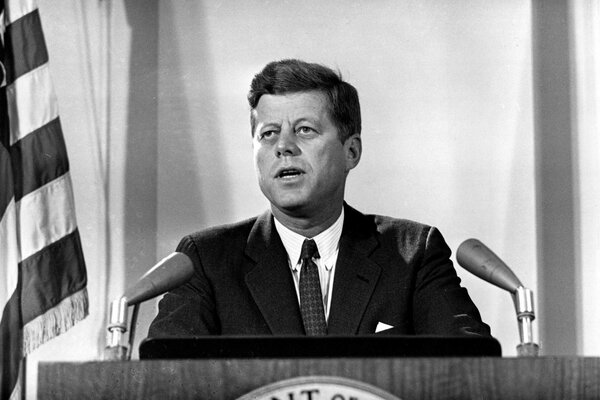 Kennedy on the podium with two microphones and a flag