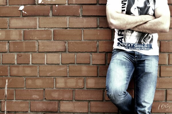 The guy is standing in jeans near a brick wall and posing