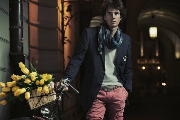 A dapper guy in stylish clothes is standing in an alley with a bicycle with a basket of yellow tulips