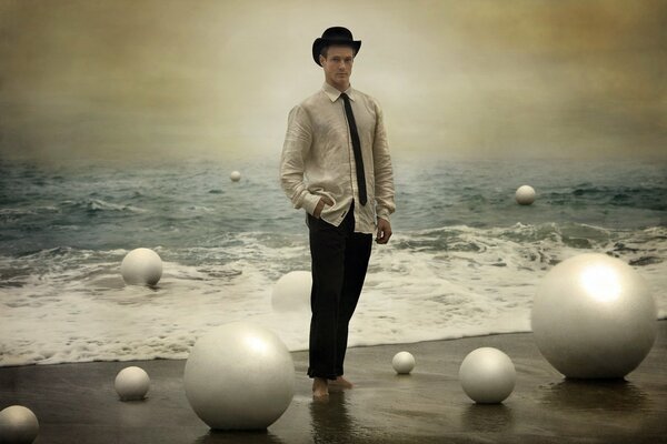 A young man in a black hat, white shirt, black tie and trousers walks barefoot along the seashore among mother-of-pearl balls