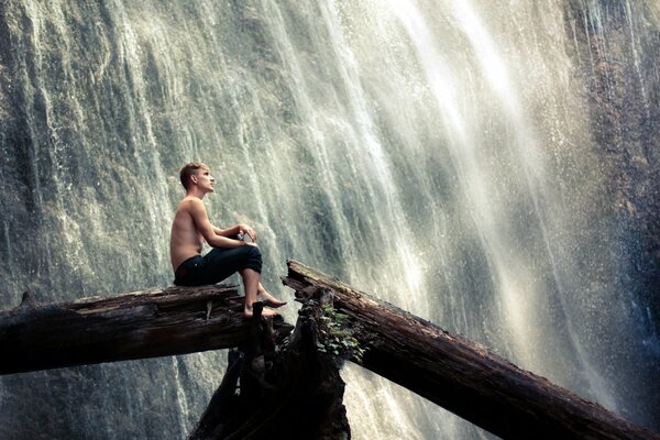 The guy is sitting on a tree by the waterfall