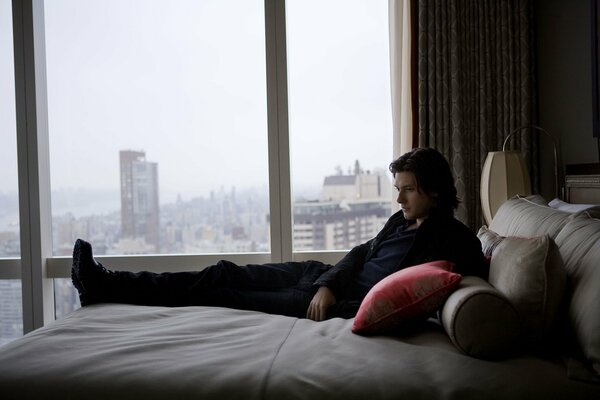 Papel pintado con Ben Barnes en la cama con almohadas. Ciudad fuera de la ventana