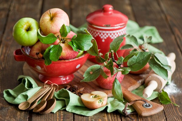 Auf einem Holzbrett Obst in einer Obstschale, Nudelholz, Pfanne, Holzlöffel und Äpfel mit Blättern