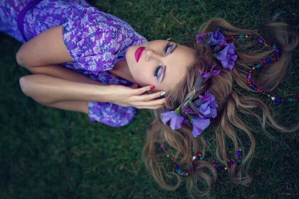 Una chica con un vestido morado y flores en el pelo yace sobre la hierba