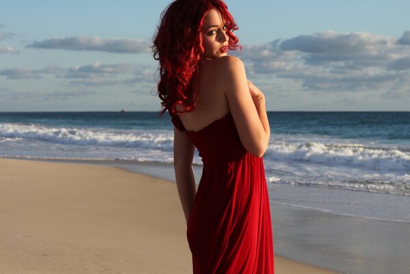A girl with red hair and a red dress stands against the background of waves