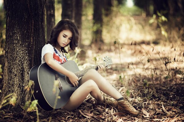 Una chica toca la guitarra apoyada contra un árbol