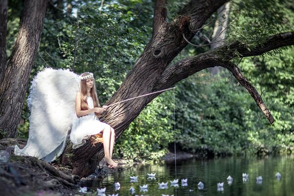 Mädchen in Gestalt eines Engels mit weißen Flügeln sitzt auf einem Baum mit einer Angelrute über einem Teich Sommerlandschaft