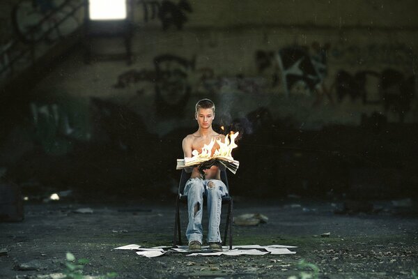 The guy is sitting on a chair with a burning newspaper