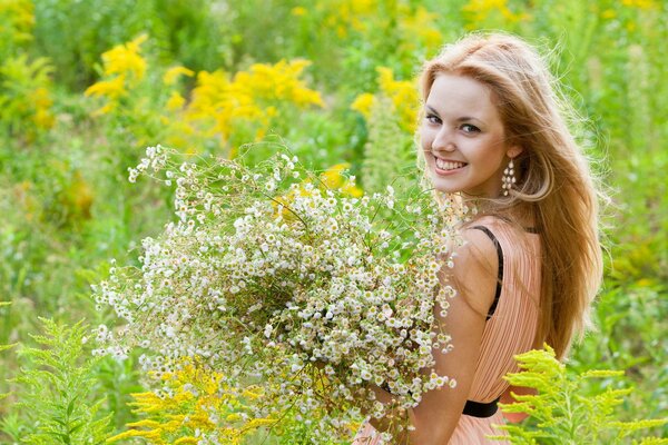 Una chica sonriente con pendientes largos se para con un ramo de flores silvestres blancas en un vestido rosa en medio de la vegetación