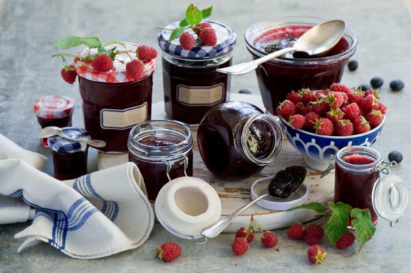 Auf dem Tisch liegen Marmeladengläser und ein tiefer Teller mit Himbeerbeeren