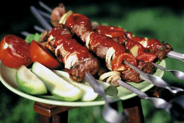 Shish kebab on skewers with tomato paste lies on a plate next to slices of tomatoes and cucumbers