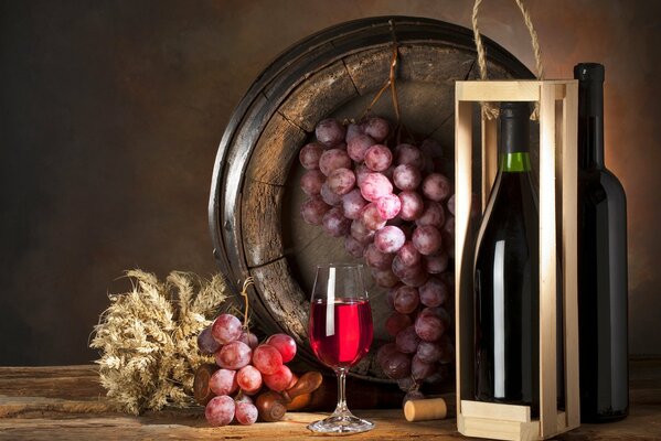 There is a bottle in a box and a glass of wine on the table, and a bunch of grapes in a barrel next to it