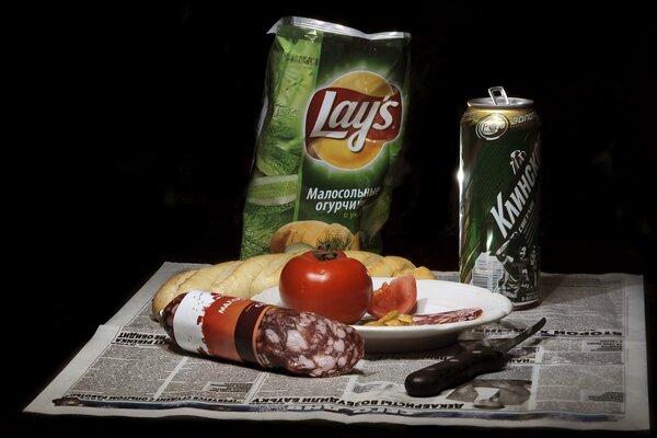 Chips, beer, tomato, loaf and sausage with a knife on the newspaper