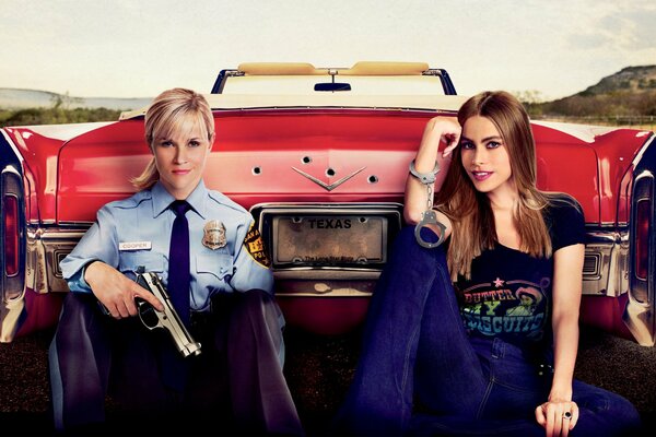 Reese Witherspoon en uniforme de police avec un pistolet et Sofia Vergara menottées sur fond de voiture rouge