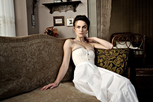 Keira Knightley in a white dress is sitting on the couch