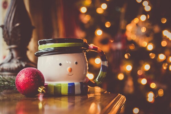 Taza-muñeco de nieve y bola de árbol de Navidad rojo en camode en el fondo de las luces