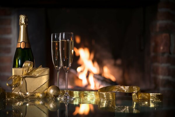 There is a bottle of champagne, glasses and a gift on the table. A fireplace is burning in the background