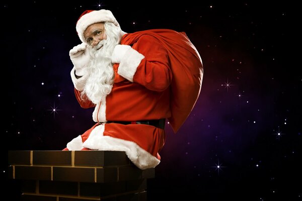 Greeting waving and smiling Santa Claus with a bag of gifts is sitting on the chimney