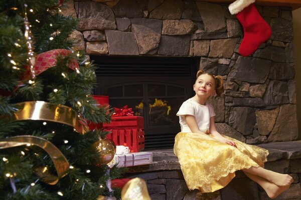 A little girl in a yellow skirt is sitting by the fireplace