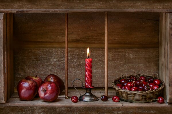 A red twisted candle burns among cherries in a basket and apples on a wooden shelf