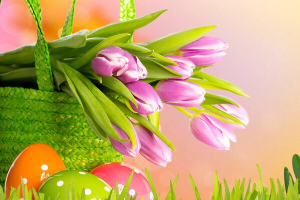 Easter eggs are lying on the green grass next to a basket of flowers