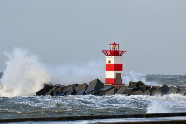 Una ola de mar se rompe contra una base rocosa con un faro rojo y blanco