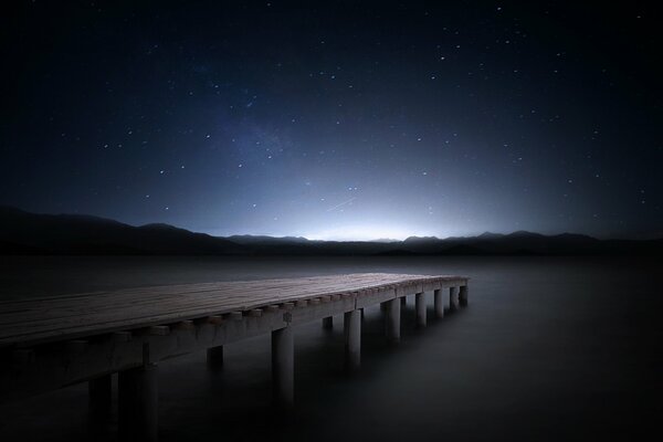 Cielo estrellado sobre el muelle cerca del lago. A lo lejos se ven las montañas